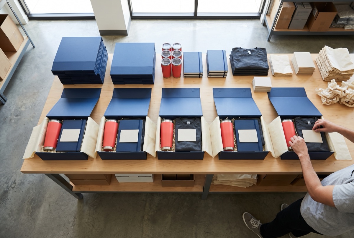 Rows of navy gift boxes being packed with coral tumblers, notebooks and t-shirts on a worktable.